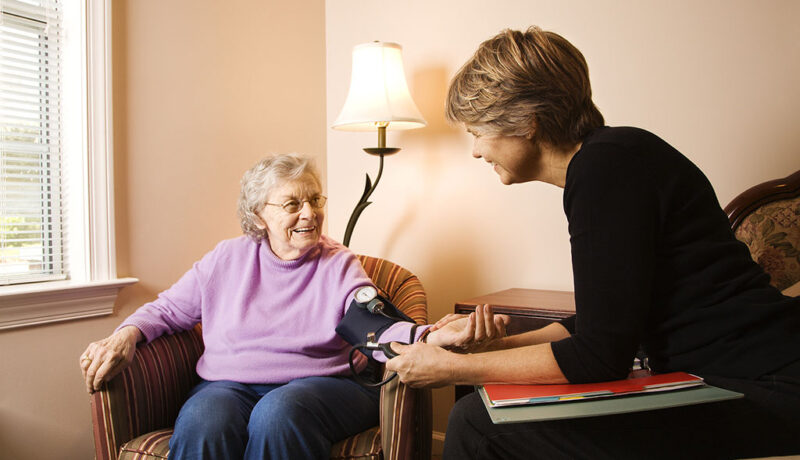 A Nurse Sitting Beside a Senior Woman’s Checking Her Blood Pressure What Are Chronic Illnesses