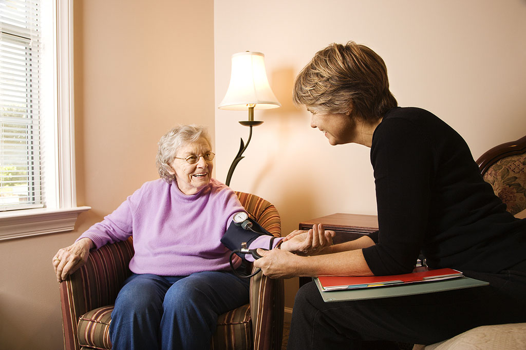 A Nurse Sitting Beside a Senior Woman’s Checking Her Blood Pressure What Are Chronic Illnesses