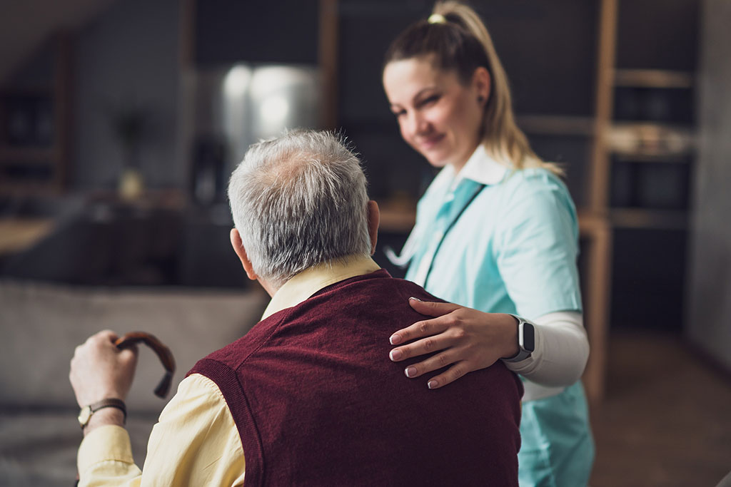 A Caregiver Smiling With Her Hand on a Senior Man’s Back Sitting Down Family Owned Assisted Living