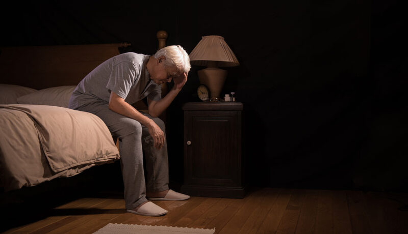 A Senior Man Sitting On The Edge Of The Bed Holding His Head Assisted Living For Sundown Syndrome