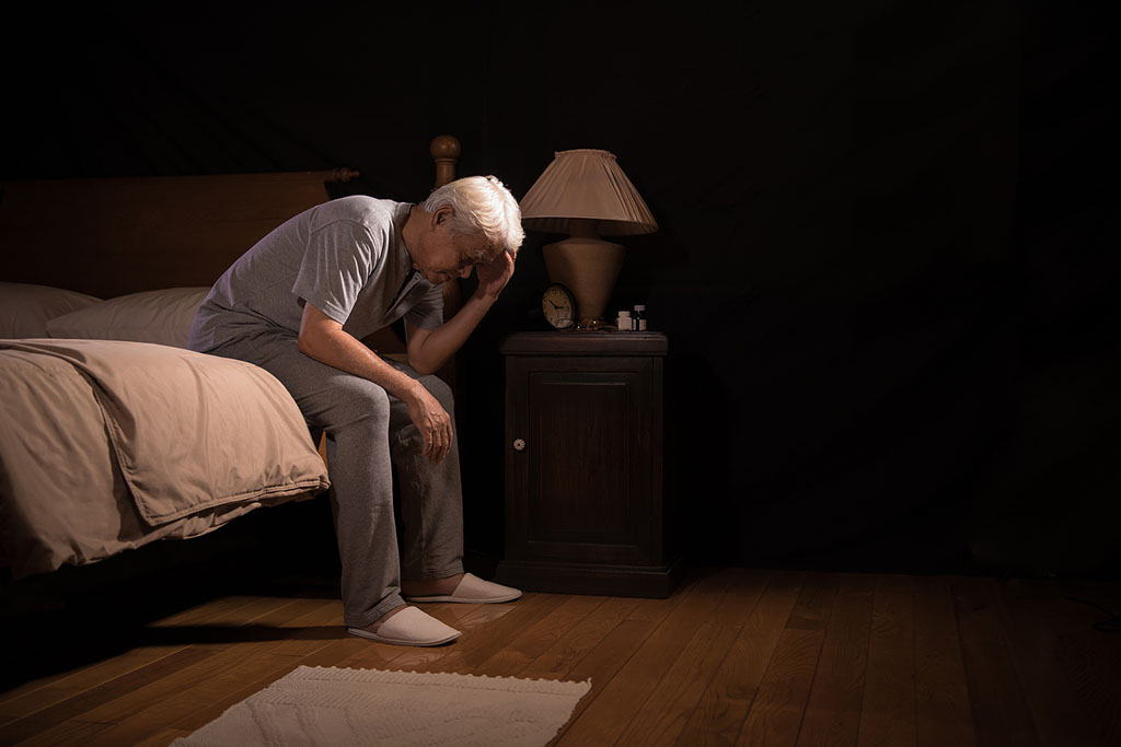 A Senior Man Sitting On The Edge Of The Bed Holding His Head Assisted Living For Sundown Syndrome